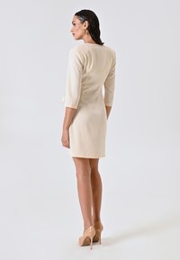 Cremefarbenes, knielanges Kleid mit Dreiviertelärmeln, glatter Textur und taillierter Passform. Kombiniert mit nudefarbenen High Heels.