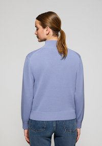 Ljuslila stickad tröja med hög krage, ribbade ärmslut och fåll. Har en slät yta och en figursydd form, bärs med blå jeans.