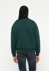 Felpa verde con scollo rotondo e polsini a costine, abbinata a jeans blu scuro. La felpa ha una vestibilità rilassata e una texture liscia.