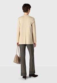 Femme debout de dos, portant un blazer beige, un pantalon à motifs, des talons noirs et tenant un sac à main beige dans la main gauche.