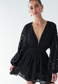 Juoda permatoma suknelė, turinti gilų V formos iškirpimą, ilgomis rankovėmis, elastiniu liemeniu ir tekstūruotą gyvūnų raštą. Papildyta juosta juosmens srityje.