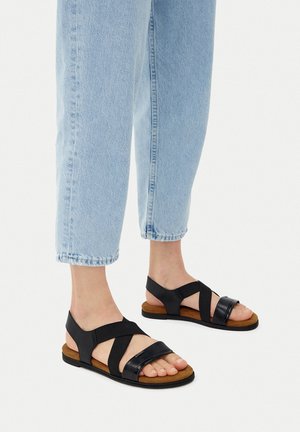 Saboți negre cu curele încrucișate, cu un finisaj lucios și neted pe partea superioară și o talpă texturată de culoare maronie. Purtați cu blugi cropped de un albastru deschis.