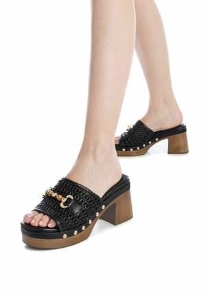 Sandalias deslizantes negras con textura trenzada, acentos en tono dorado, tacones cuadrados marrones gruesos y tachuelas decorativas a lo largo de la suela.