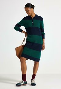 Stickad klänning med gröna och marinblå horisontella ränder, polo-krage, knäppning framtill, lång ärm, kombinerad med en brun väska och marinblå sneakers.