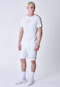 Conjunto de camiseta de manga corta blanca y pantalones cortos, con letras del logo en dorado y franjas laterales en negro. Fabricado con tejido suave, incluye zapatillas blancas.