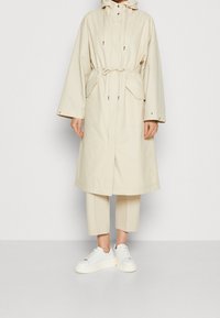 Cappotto impermeabile beige con cappuccio regolabile, tasche frontali e vita regolabile, abbinato a pantaloni cropped crema e sneakers bianche.