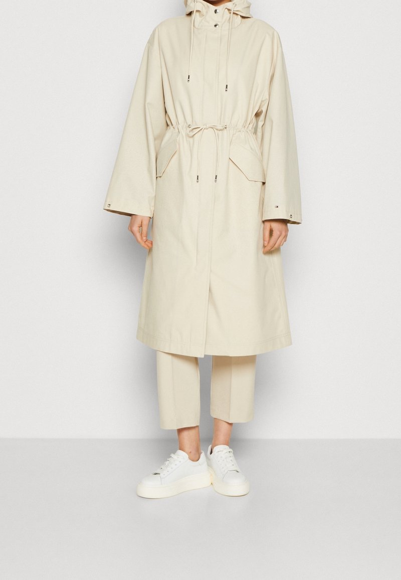 Cappotto impermeabile beige con cappuccio regolabile, tasche frontali e vita regolabile, abbinato a pantaloni cropped crema e sneakers bianche.