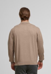 Man met bruin haar in een knot, gekleed in een beige long-sleeve top en donkere grijze broek, staand met de rug naar de camera tegen een effen achtergrond.