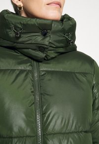 Doudoune verte fermée avec col montant, zip noir et boutons-pression, portée par une personne avec des boucles d'oreilles créoles dorées.