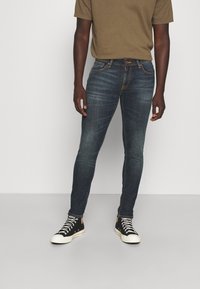 Mörkblå skinny jeans med stickade fickor, guldfärgad beslag och en lätt blekning, matchade med en brun kortärmad skjorta och svarta sneakers.