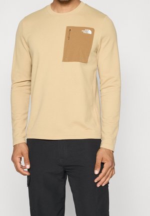 Mann trägt beige langärmeliges Hemd mit strukturierter Stoffoberfläche und einer hellbraunen Brusttasche mit Reißverschluss, auf der das The North Face-Logo zu sehen ist, kombiniert mit einer schwarzen Hose.