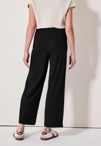 Zwarte wijde broek met een elastische tailleband, twee achterzakken, gedragen met witte sneakers met rode accenten en een bruine zool.