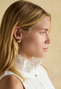 Zlaté kruhové náušnice zdobí ucho ženy, která má na sobě bílou volánkovou límeček. Pozadí je v jemném, neutrálním tónu. Vlasy jsou rovné a blond.