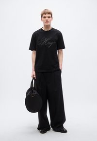 Μαύρο t-shirt με λευκό λογότυπο «Hugo», συνδυασμένο με φαρδιά μαύρη παντελόνια. Το μοντέλο κρατάει μια στρογγυλή μαύρη τσάντα με χειρολαβή στην κορυφή.