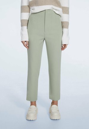 Persona con pantalones ajustados verde claro, suéter a rayas beige y blanco, y mocasines blancos gruesos con plataforma, de pie sobre un fondo claro.