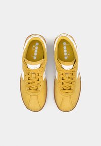 Yellow suede sneakers with white accents, round toe, and flat laces. Features textured fabric and a rubber sole. Logo on the tongue.