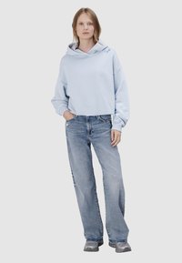 Kobieta stojąca z jedną ręką w kieszeni, ubrana w jasnoniebieską krótką bluzę z kapturem, luźne wyblakłe dżinsy i szare tenisówki na jednolitym tle.