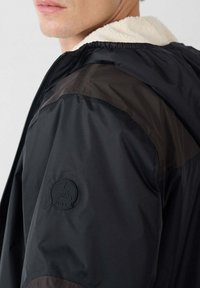 Chaqueta negra con un forro suave de color crema. Cuenta con cierre de cremallera, grandes secciones en los hombros y un parche circular con logo en el brazo.