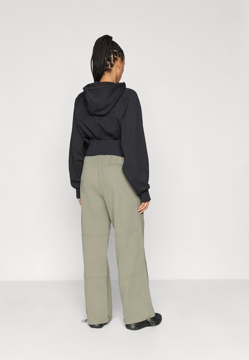 Vrouw met gevlochten haar, gekleed in een zwarte cropped hoodie en losse lichtgrijze broek, staande met haar gezicht naar een witte muur.