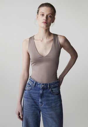 Donna con capelli biondi, indossa un top senza maniche taupe e jeans blu a vita alta, in piedi con una mano sui fianchi davanti a uno sfondo chiaro e uniforme.