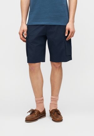 Uomo con pantaloncini cargo blu navy, calzini pesca e scarpe da barca in pelle marrone, in piedi davanti a uno sfondo bianco.