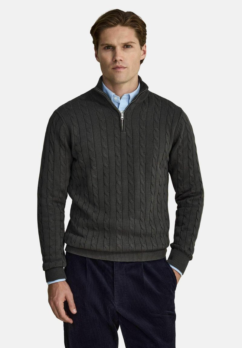 Homme portant un pull gris foncé à col zippé en maille torsadée par-dessus une chemise bleu clair à boutons et un pantalon foncé, debout avec une main dans la poche.