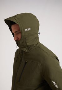 Oliven grøn jakke med hætte, der har en fuld lynlås fortil, snøre detaljer og en frontlomme. "JACKIT" logo trykt i hvid.