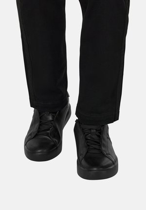 Sorte lædersneakers med snørebånd iført med sorte bukser foran en ensfarvet lys baggrund, fødderne peger fremad.