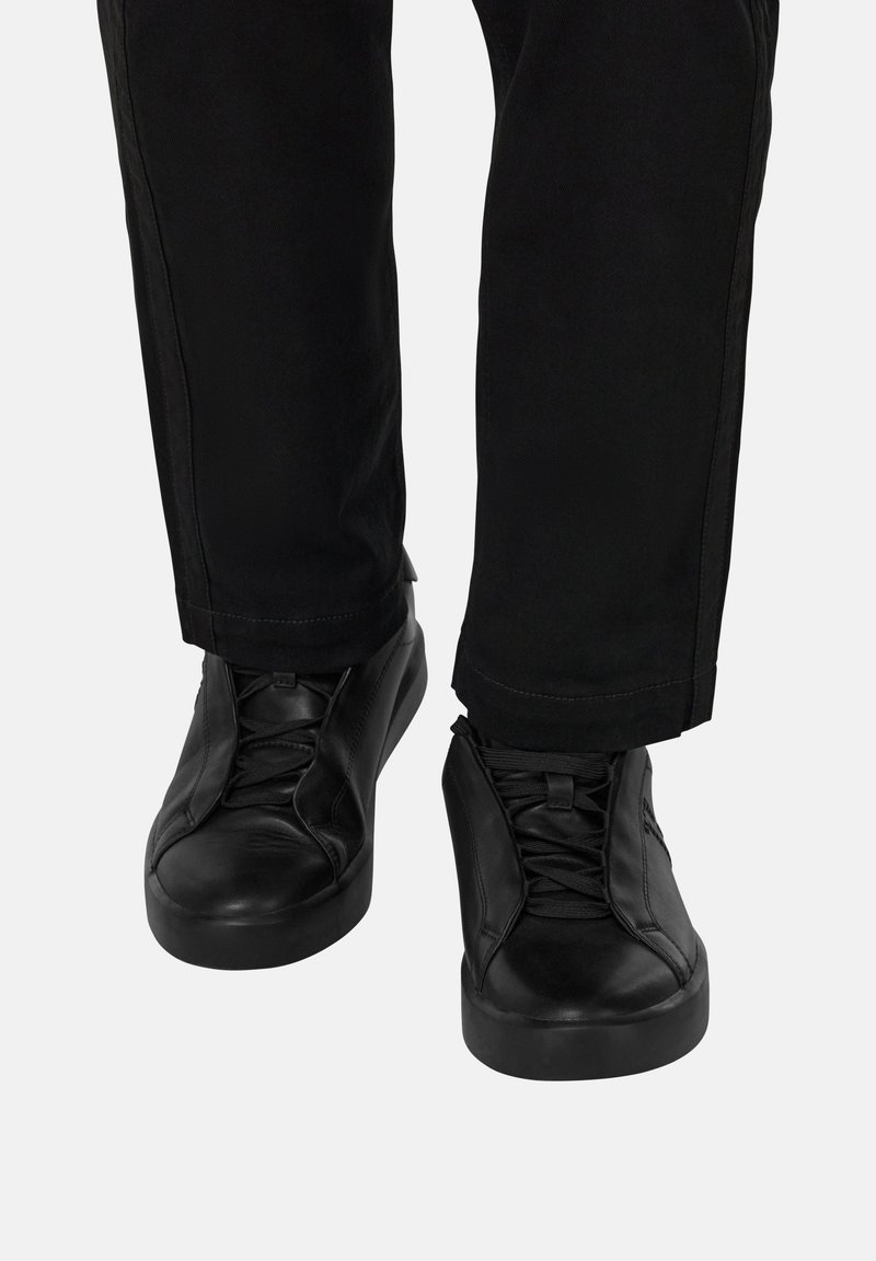 Czarne skórzane sneakersy z sznurówkami noszone z czarnymi spodniami na jednolitym jasnym tle, stopy skierowane do przodu.