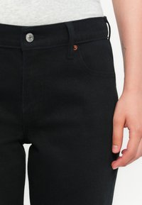 Nærbilde av en person som har på seg svarte jeans med sølvknapp, med en synlig hånd hvilende ved siden av låret.