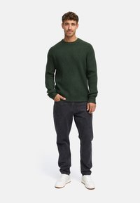 Suéter verde con relieve y escote redondo, combinado con jeans de mezclilla oscuros y zapatillas blancas. La tela parece texturizada y cálida.