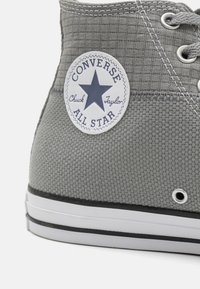 Close-up of gray Converse All Star Chuck Taylor sneaker showing textured fabric, white rubber sole, metal eyelets, and logo patch.