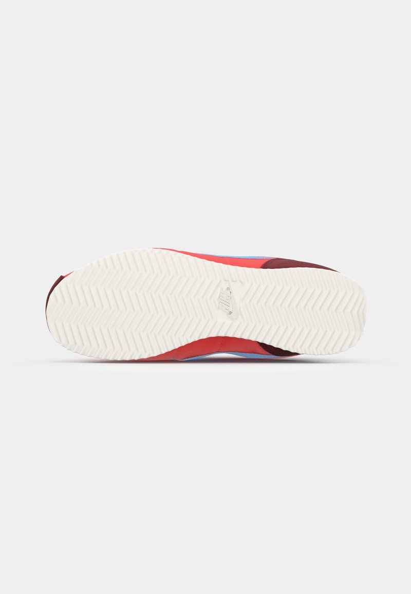 Zapatilla roja y azul con una suela de goma blanca texturizada. La parte superior presenta una tela suave con un diseño estampado y una sutil marca.