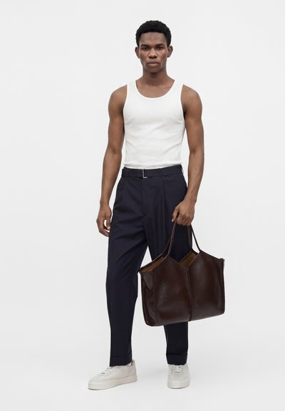 Jeune homme portant un débardeur blanc, un pantalon bleu marine, des baskets blanches, tenant un grand sac cabas en cuir marron foncé dans la main droite.