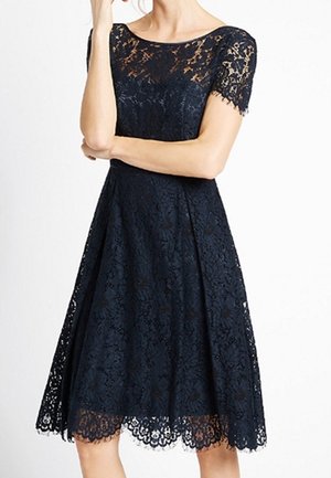 Frau in einem knielangen, kurzärmligen, marineblauen Spitzenkleid mit Blumenmustern und gezacktem Saum, stehend mit verschränkten Armen.