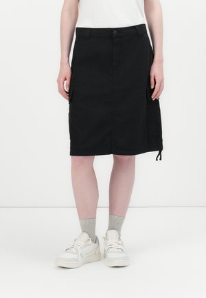 Vrouw draagt een zwarte knielange rok met zijzakken, grijze sokken, witte sneakers en een wit topje tegen een effen witte achtergrond.