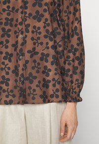 Blusa marrone con motivo floreale nero, chiusura a bottoni e polsini arricciati. Tessuto leggero che cade morbidamente, completando i pantaloni beige.