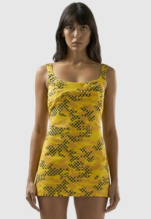 Mujer con un vestido mini amarillo sin mangas con patrones de cuadros negros y camuflaje, de pie sobre un fondo liso gris claro.