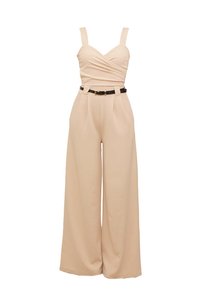 Combinaison beige avec un corsage ajusté, design croisé, pantalon évasé et une ceinture noire avec une boucle en or. Texture de tissu lisse.