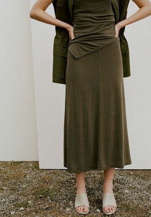 Frau trägt olivgrünes ärmelloses Kleid mit passendem Blazer über den Schultern, steht auf Kies und trägt beige offene Mules.