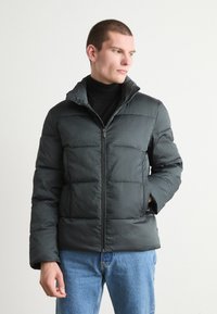 Giacca puffer verde scuro con texture trapuntata, colletto alto, zip frontale completa e tasche laterali, indossata sopra un dolcevita nero.