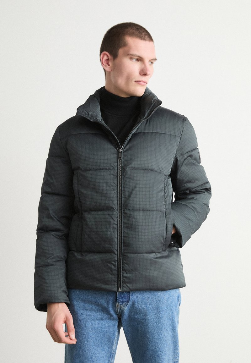 Giacca puffer verde scuro con texture trapuntata, colletto alto, zip frontale completa e tasche laterali, indossata sopra un dolcevita nero.