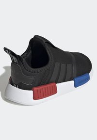 Svart sportskor med en slank design, som har tre svarta ränder, en texturerad ovandel och röda samt blå detaljer på den vita sulan.