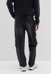 Zwarte cargobroek met een losse pasvorm, voorzien van meerdere zakken en zichtbare stiksels. Draagt witte sneakers en een donkere trui.