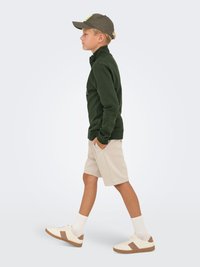 Grøn lynlås sweatshirt, beige shorts, hvide sneakers med brune detaljer, grå kasket. Barn går i profil med hænderne i lommerne.