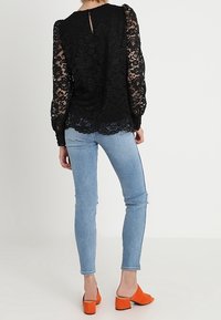Top in pizzo nero con maniche lunghe, caratterizzato da un dettaglio a chiave sul retro. Abbinato a jeans skinny azzurri chiari e mule con tacco a blocco arancioni.