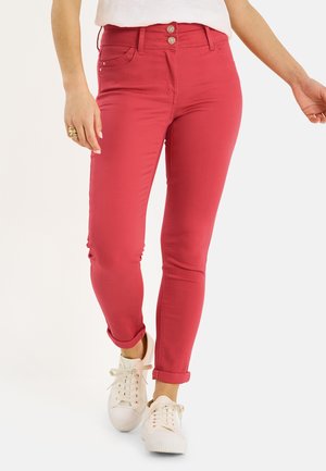 Mujer con pantalones pitillo rojos remangados y zapatillas blancas, de pie con un pie ligeramente adelantado sobre un fondo blanco.