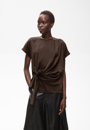 Femme aux cheveux courts portant un haut marron foncé à manches courtes avec un nœud devant et un pantalon large en cuir noir, debout sur fond blanc.