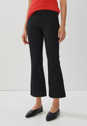 Frau in schwarzen, cropped Schlaghosen, schwarzen spitzen Ballerinas und einem roten ärmellosen Oberteil, die vor einem schlichten weißen Hintergrund steht.