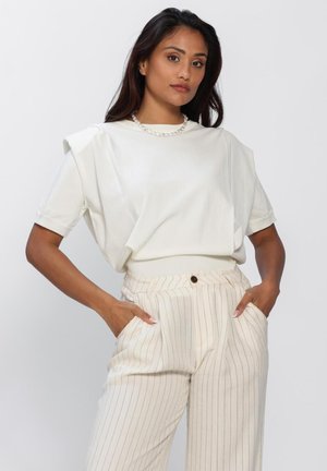 Vrouw met lang, donker haar, gekleed in een wit topje met gewatteerde schouders, een parelketting en crèmekleurige, hooggetailleerde broek met krijtstreepjes, met haar handen in haar zakken.